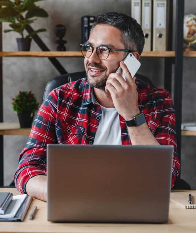 Man in front of his laptop speaking on a cell phone.